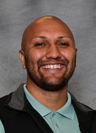 Professional headshot of smiling man with beard wearing light blue shirt.