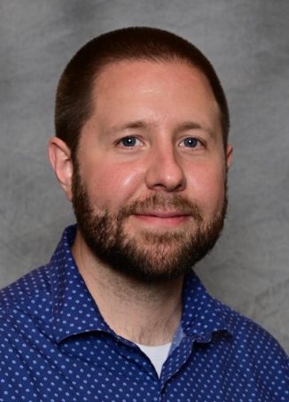 Professional headshot of bearded man in blue patterned shirt.