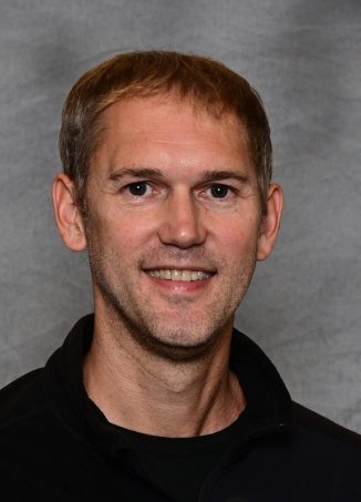 Professional headshot of man with brown hair wearing black shirt.