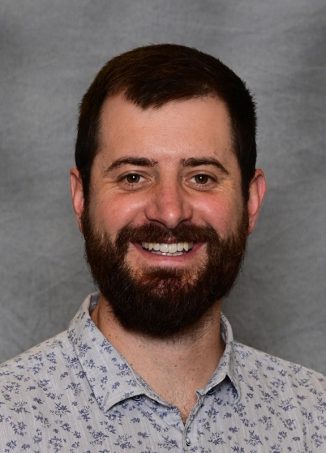 Man with dark beard wearing patterned blue shirt smiling.