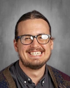 Man wearing glasses and dark collared shirt, smiling.